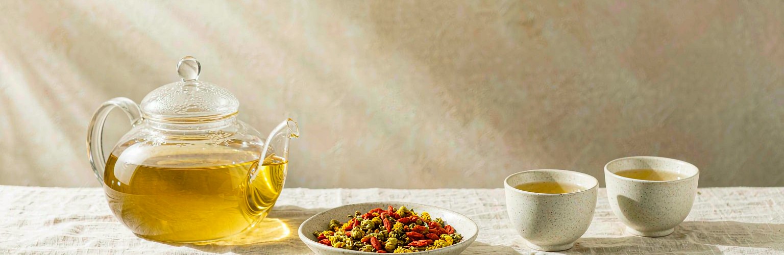 A bright still life on warm linen: a glass teapot of pale herbal tea, two small ceramic cups, and a shallow dish of dried chrysanthemum and goji berries, lit by soft morning light.